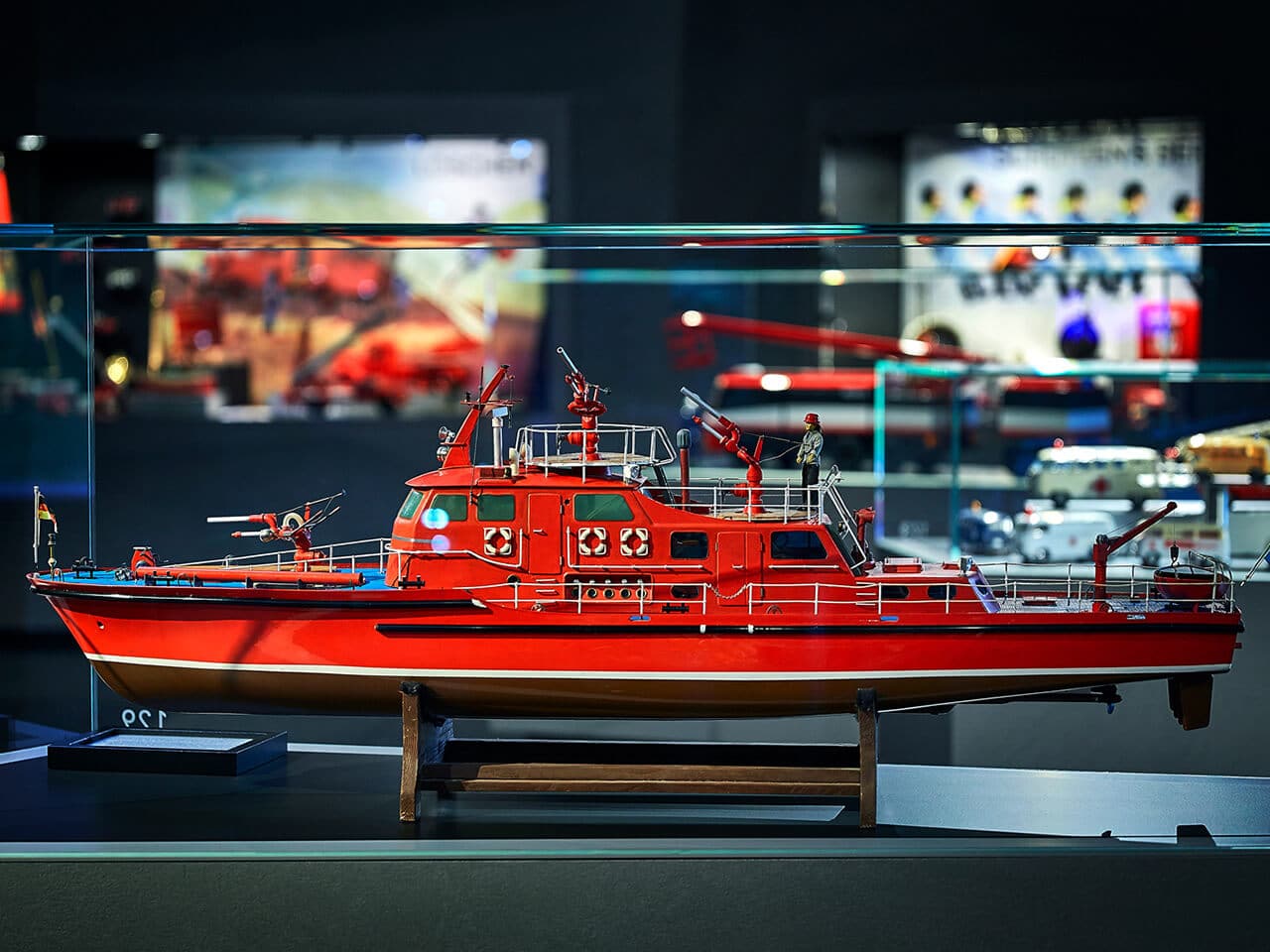 Ein detailliertes Modell eines leuchtend roten Feuerlöschboots ist in einer Glasvitrine im Traumwerk ausgestellt, mit Feuerlöschgeräten und Miniaturfiguren der Besatzung an Deck. Im Hintergrund sind weitere maritime Exponate zu sehen, was es zu einem idealen Ausflugsziel für Enthusiasten macht.