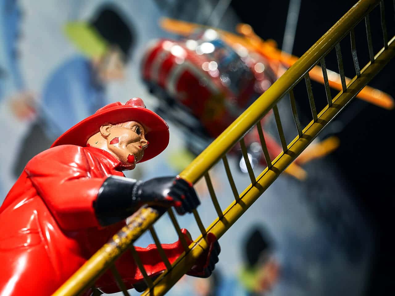 Nahaufnahme eines Feuerwehrmanns in roter Uniform, der eine gelbe Leiter hochklettert, mit einem Spielzeug-Porsche-Hubschrauber im Hintergrund.