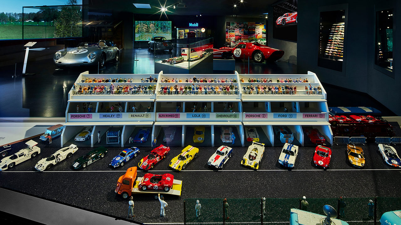 Detailgetreues Modell eines Boxenstopps eines Oldtimer-Rennens im Traumwerk mit Miniatur-Porsche-Rennwagen, Boxencrews, Zuschauern und Garagen, die mit berühmten Rennmarken beschriftet sind; im Hintergrund sind echte Oldtimer zu sehen - ein perfektes Ausflugsziel.