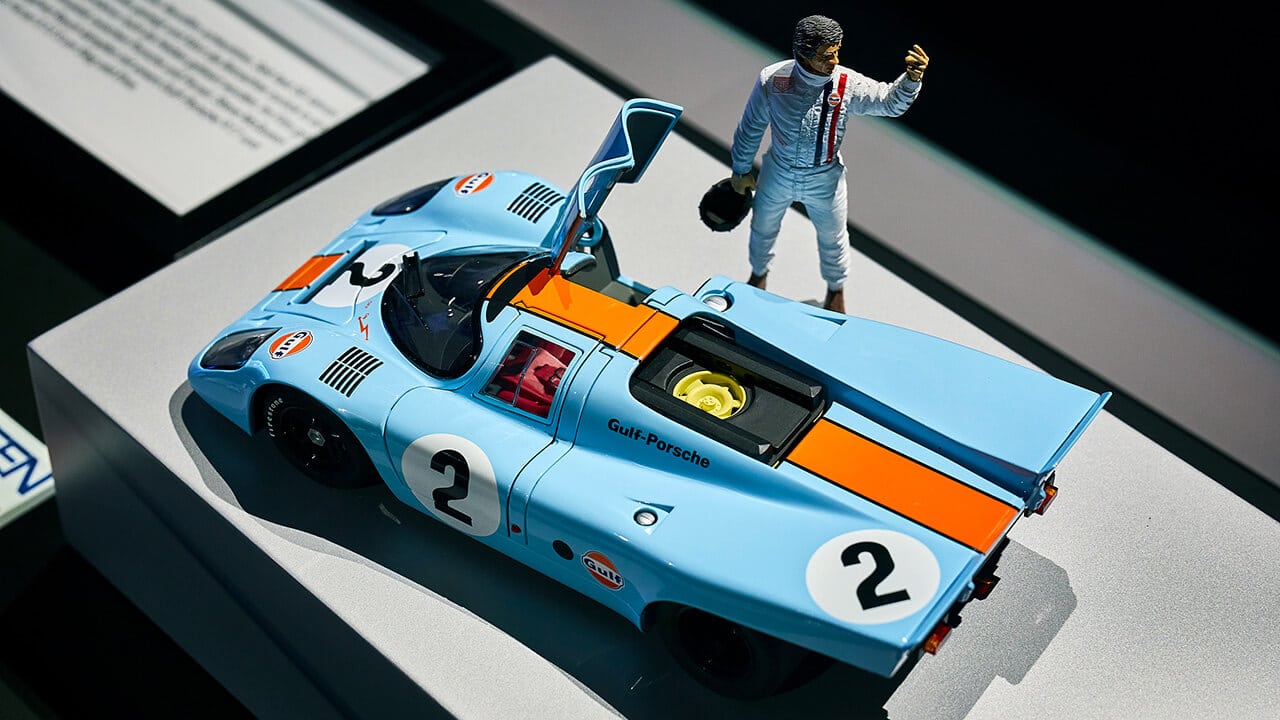 Ein Modell eines blau-orangen Porsche Gulf Rennwagens mit der Nummer 2, dargestellt mit einer kleinen Figur eines Fahrers im weißen Rennanzug mit Helm, der neben dem Auto steht - ein perfektes Highlight für jedes Traumwerk-Ausflugsziel.