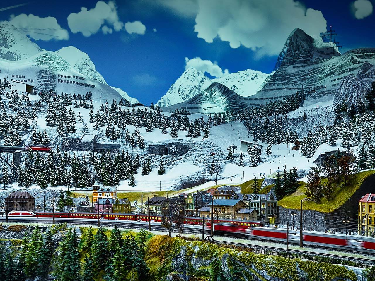Eine detaillierte Modelleisenbahnanlage im Traumwerk zeigt rote Züge, die durch eine verschneite Berglandschaft mit Tannenbäumen, kleinen bunten Gebäuden und dramatischen, schneebedeckten Gipfeln unter einem teilweise bewölkten Himmel fahren.