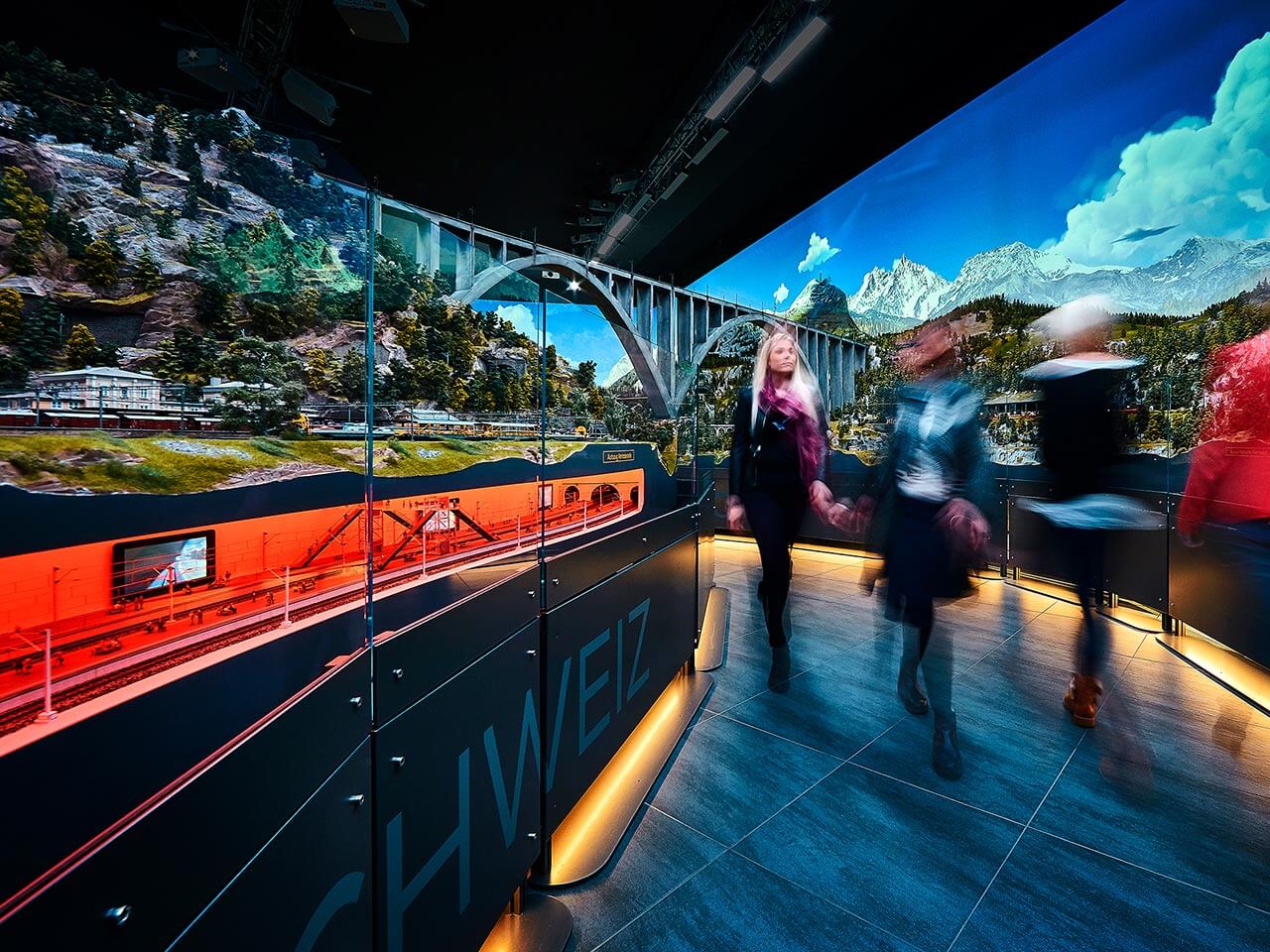 Drei Personen gehen an einer detaillierten Modelleisenbahn- und Landschaftsausstellung im Traumwerk vorbei, die eine Brücke, eine Berglandschaft und helle digitale Bildschirme zeigt, während SCHWEIZ auf einer Glaswand zu sehen ist - ein ideales Ausflugsziel für Enthusiasten.