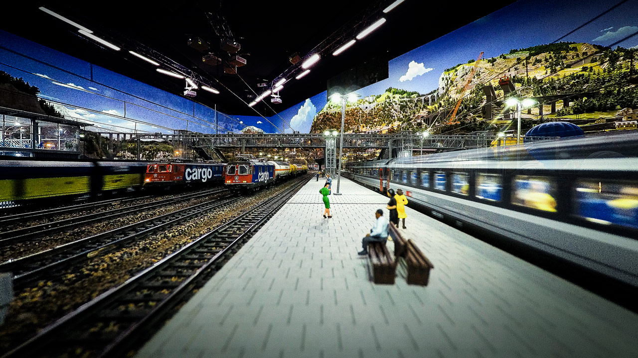 Ein detailgetreuer Modellbahnhof am Traumwerk mit Miniaturmenschen auf dem Bahnsteig, mehreren Modellzügen auf Gleisen und einer malerischen Kulisse mit Hügeln, Bäumen, Gebäuden und blauem Himmel - ein ideales Ausflugsziel für Porsche-Fans.