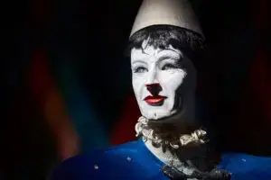 Eine als Pantomime oder Clown gekleidete Person mit weißer Gesichtsbemalung, roten Lippen und einem weißen Spitzhut, die ein blaues Kostüm trägt, wird vor einem dunklen Hintergrund dramatisch beleuchtet - wie eine Szene aus Traumwerk.