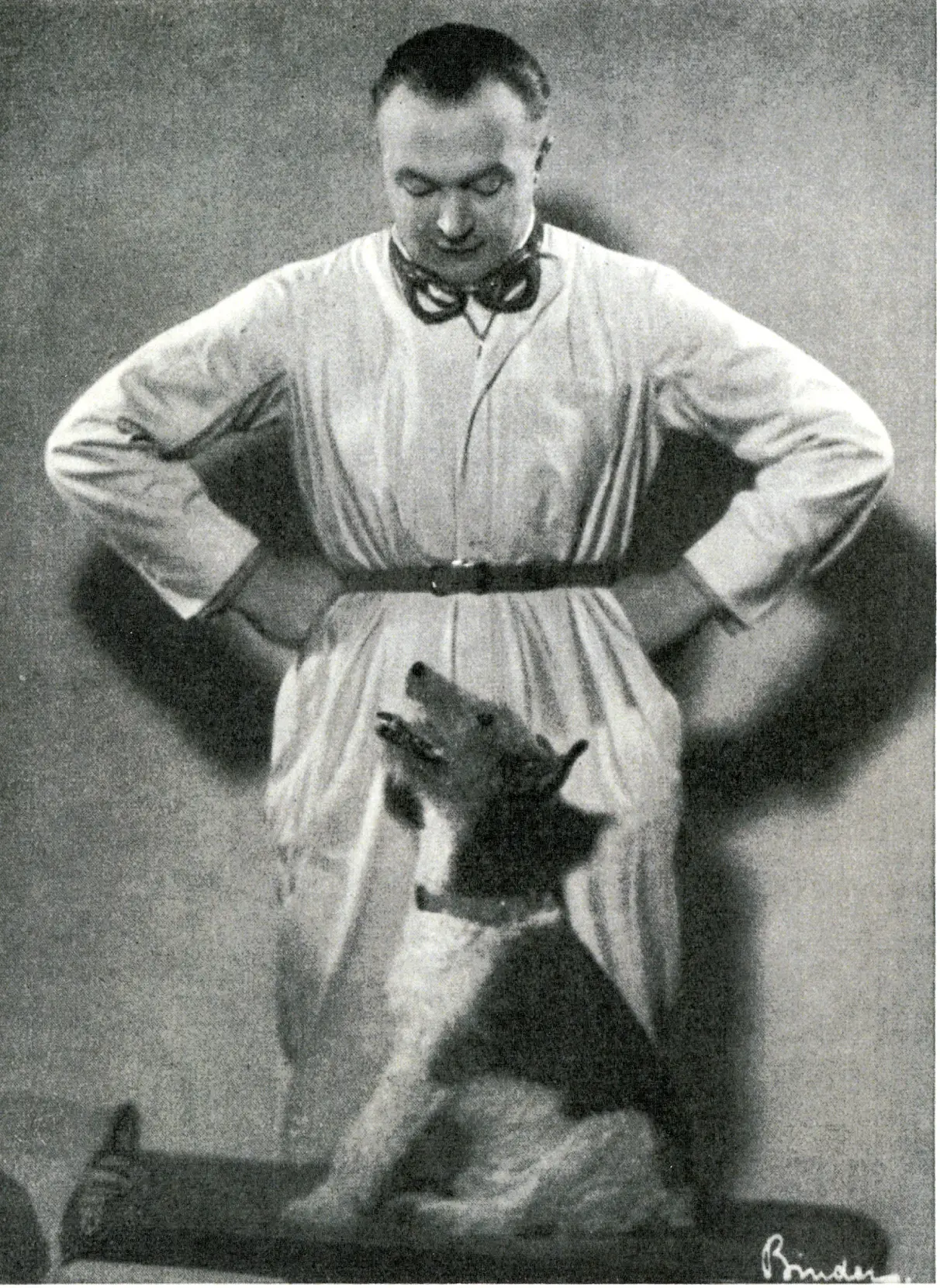 Ein Mann im Laborkittel steht mit den Händen in den Hüften da und blickt auf einen Hund hinunter, der vor ihm sitzt. Der Hund blickt zu dem Mann auf. Das Bild ist schwarz-weiß.