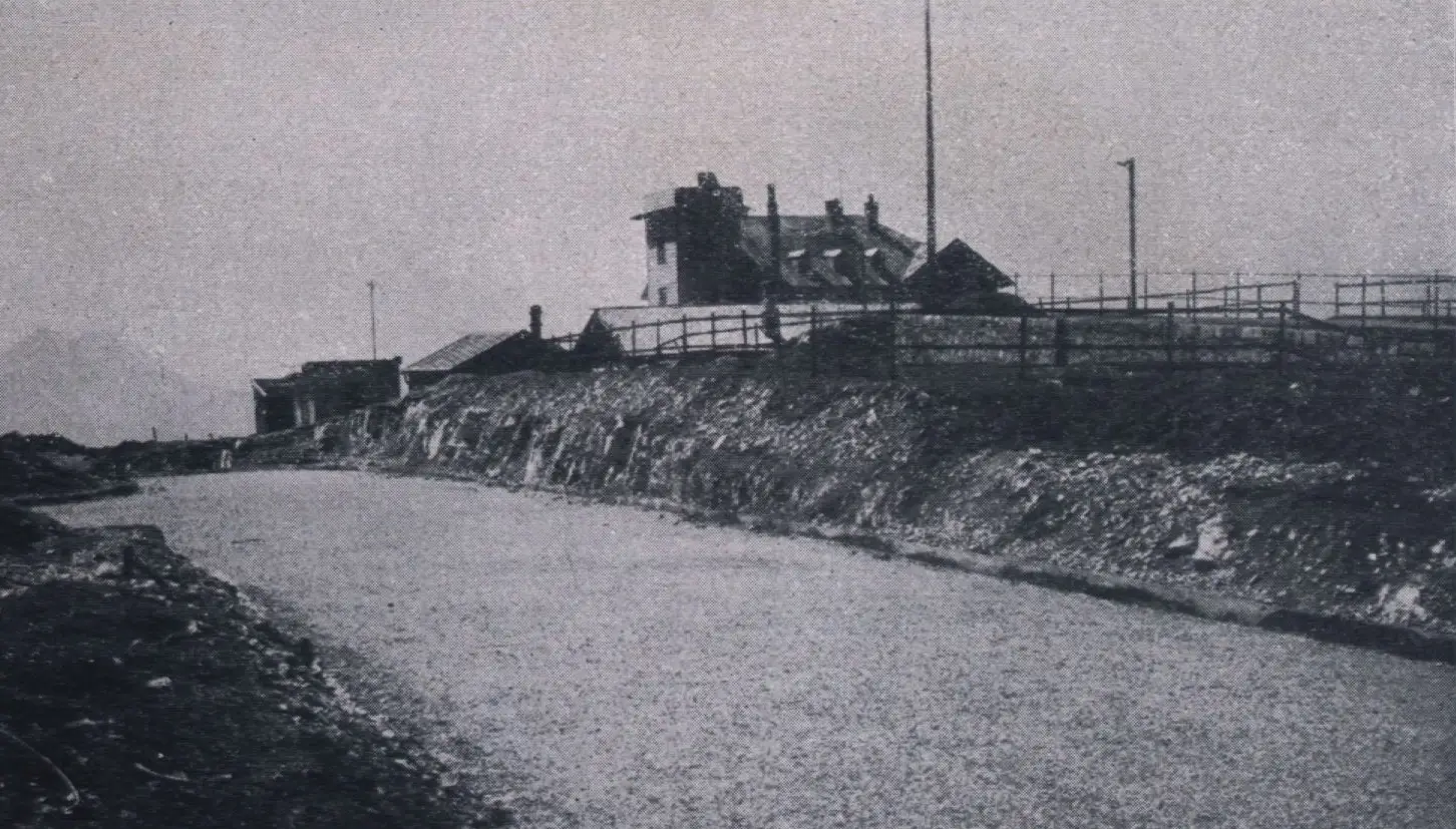 Ein Schwarzweißfoto einer verlassenen, neu asphaltierten Straße, die an einem bewölkten Tag bergauf zu einem großen Haus mit Schornsteinen führt, umgeben von Zäunen und Strommasten.