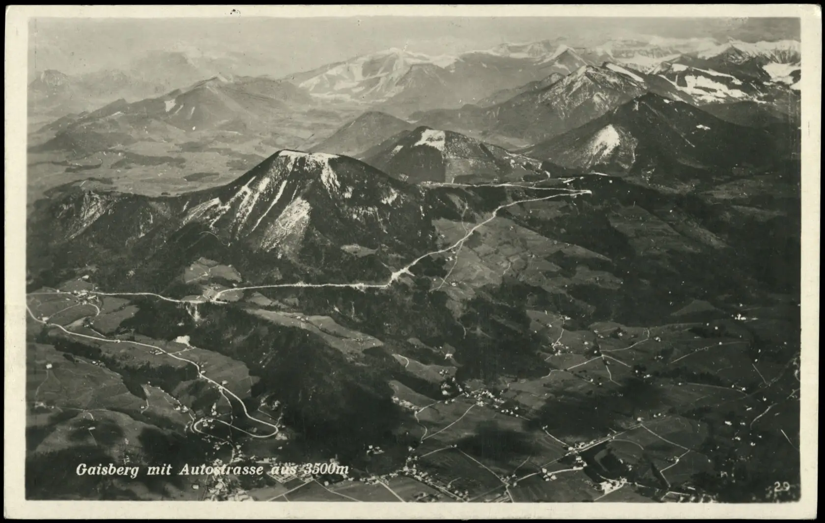 Ein Schwarz-Weiß-Luftbild zeigt kurvenreiche Straßen auf dem Gaisberg, umgeben von Tälern und schneebedeckten Gipfeln in den österreichischen Alpen. Die Landschaft ist geprägt von Wäldern, Feldern und verstreuten Gebäuden.