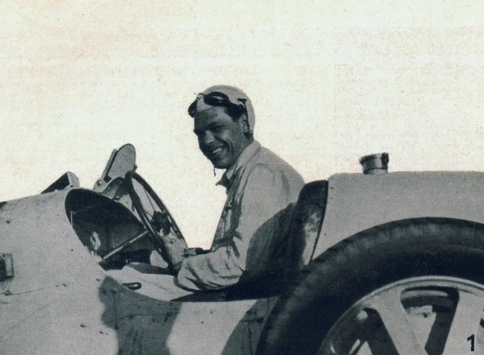 Ein altes Foto eines lächelnden Mannes mit Helm und Schutzbrille, der auf dem Fahrersitz eines alten Porsche-Rennwagens sitzt, von der Seite gesehen. Das Bild hat ein körniges, historisches Gefühl, das an frühe ausflugsziel-Abenteuer erinnert.