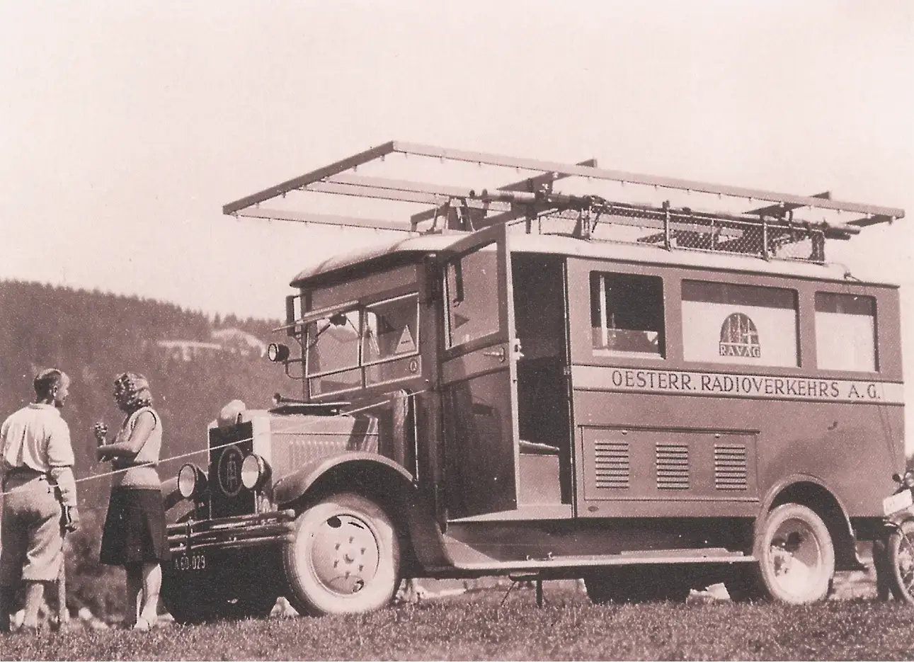 Ein alter Übertragungswagen mit Antennen auf dem Dach ist im Freien in der Nähe von Bäumen und Hügeln geparkt. Zwei Frauen und ein Mann stehen in der Nähe und unterhalten sich. Auf dem Wagen steht OESTERR. RADIOVERKEHRS A.G.-ein einzigartiges Ausflugsziel für Porsche- und Traumwerk-Liebhaber.