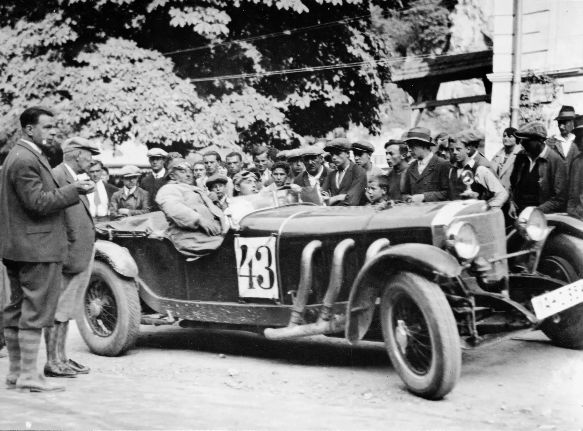 Ein Oldtimer-Rennwagen mit der Nummer 43 wird auf einer von Bäumen gesäumten Straße von einer Menschenmenge umringt, von denen einige Hüte und Anzüge tragen - ein ideales Ausflugsziel für Porsche- und Traumwerk-Liebhaber aus dem frühen 20.