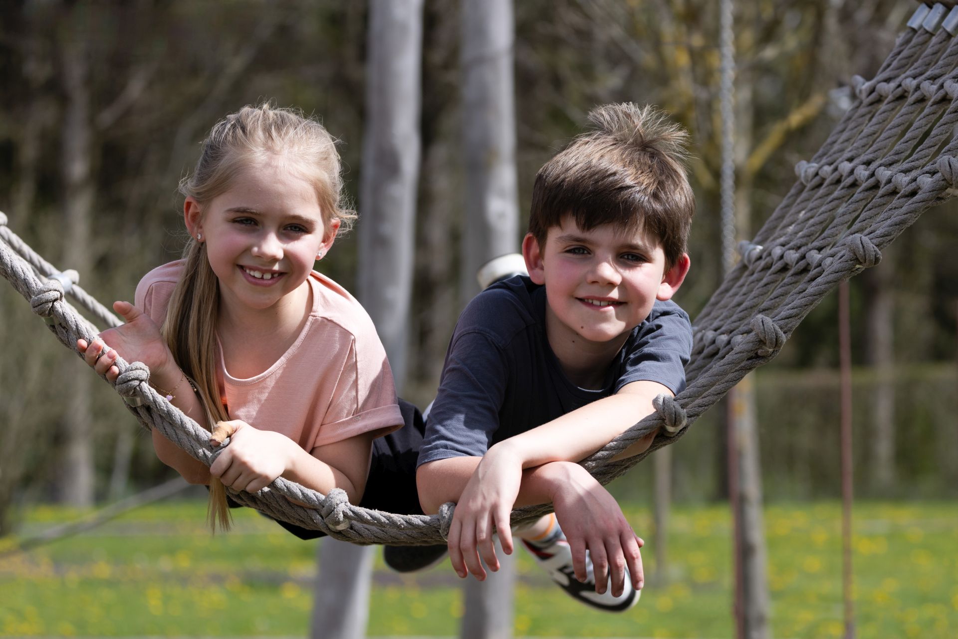 Zwei lächelnde Kinder, ein Mädchen und ein Junge, entspannen sich an einem sonnigen Tag auf einer Seilhängematte im Freien bei Traumwerk, mit grünem Gras und Bäumen im Hintergrund. Ein perfektes Ausflugsziel für Familien.