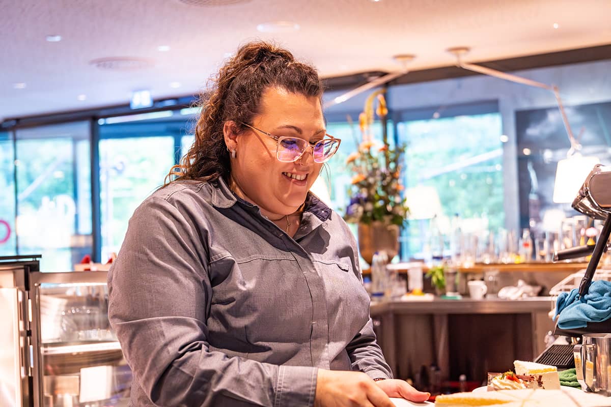 Eine Frau mit lockigem Haar und Brille lächelt, während sie hinter einer Theke in einem hell erleuchteten Café oder Restaurant Essen serviert.