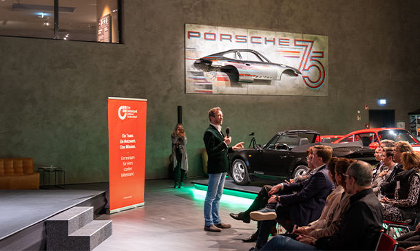 Ein Mann spricht in einem modernen Veranstaltungsort zu seinem Publikum. Porsche-Klassiker sind ausgestellt. Auf einem großen Wandbild steht „Porsche 75“. In Bühnennähe hängt ein orangefarbenes Banner. Die Leute sitzen und hören aufmerksam zu.