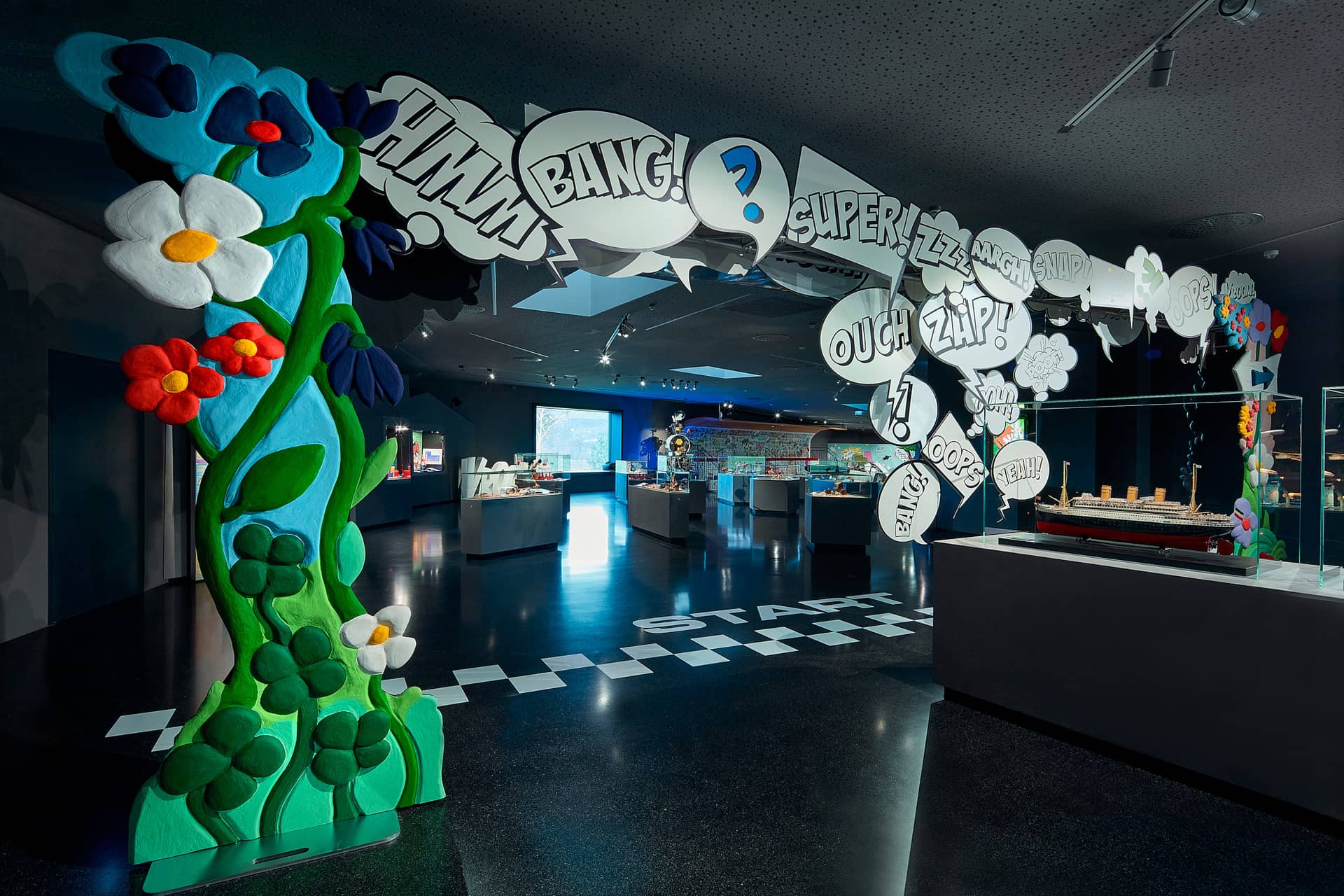 Der farbenfrohe Ausstellungseingang ist mit großen Cartoon-Blumen und Sprechblasen im Comic-Stil mit Wörtern wie BANG! und ZAP! geschmückt. Im Inneren präsentieren Vitrinen verschiedene Gegenstände unter heller Beleuchtung.