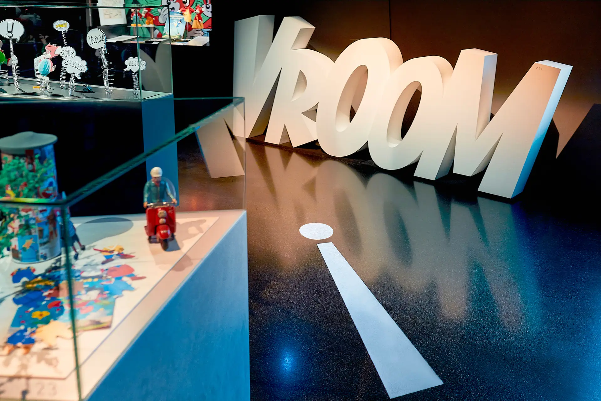 Große weiße Buchstaben mit dem Wort „VROOM“ stehen auf einem dunklen Boden neben einer weiß gemalten Linie, die einer Straße ähnelt. Im Vordergrund enthält eine Glasvitrine buntes Spielzeug, darunter einen roten Roller mit einer kleinen Figur.