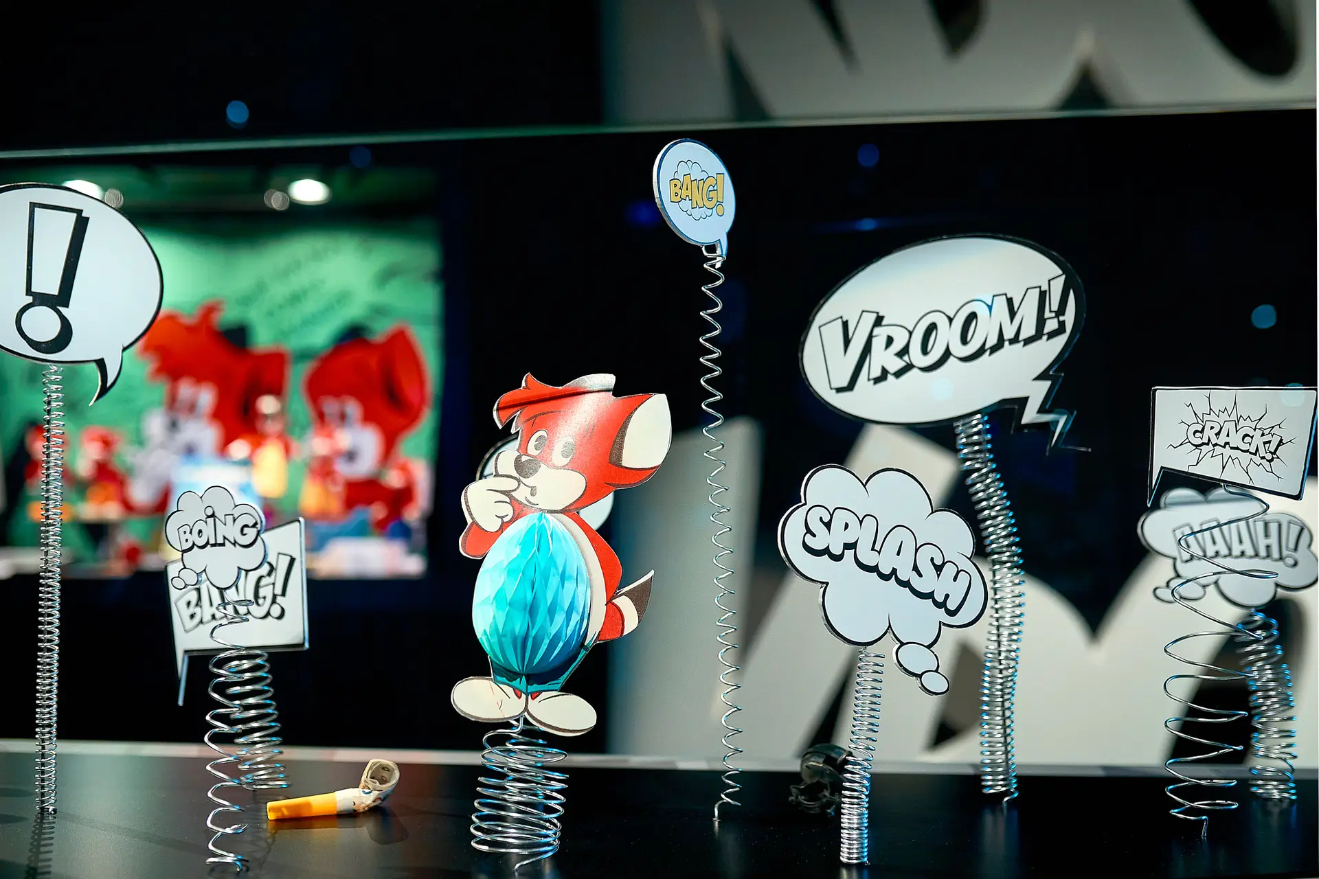 Comic-Display mit einem Zeichentricktier, Sprechblasen mit den Worten „VROOM!“ und „SPLASH!“ sowie an Schildern befestigten Federn. Auf der Oberfläche liegt ein gelbes Horn, im Hintergrund ist buntes Zeichentrick-Design zu sehen.