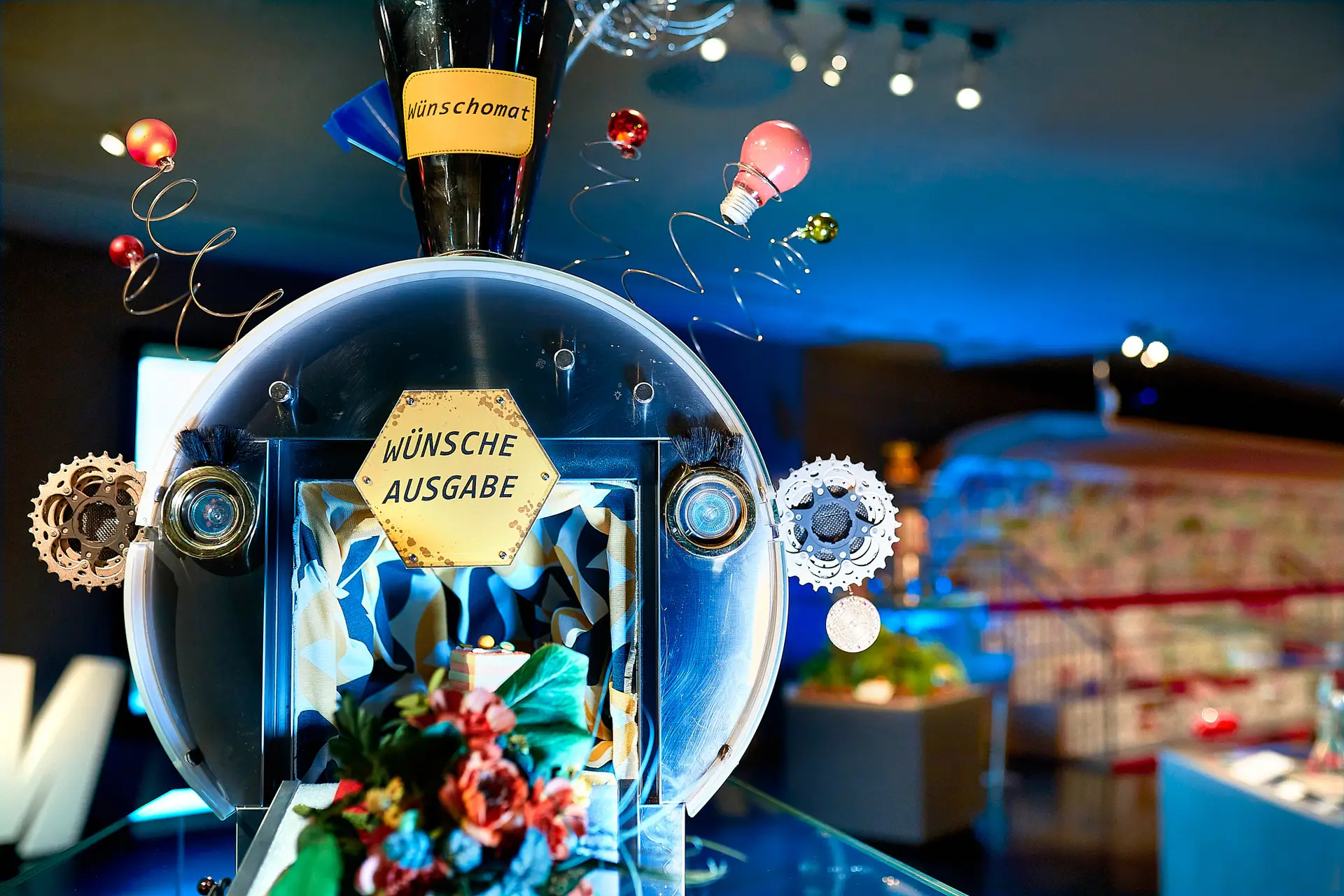 Eine skurrile Maschine mit Zahnrädern, Glühbirnen und einem Schild mit der Aufschrift „Wünsche Ausgabe“ steht unter hellem Licht, dekoriert mit bunten Blumen und metallischen Spiralen in einem modernen, kreativen Ambiente.