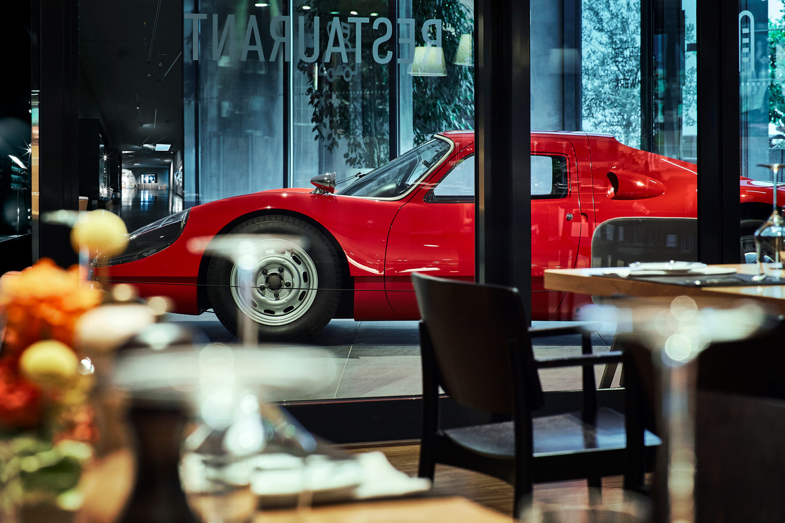 Vor einem modernen Restaurant steht hinter Glas ein klassischer roter Sportwagen, im Vordergrund sind verschwommene Esstische und Stühle zu sehen.