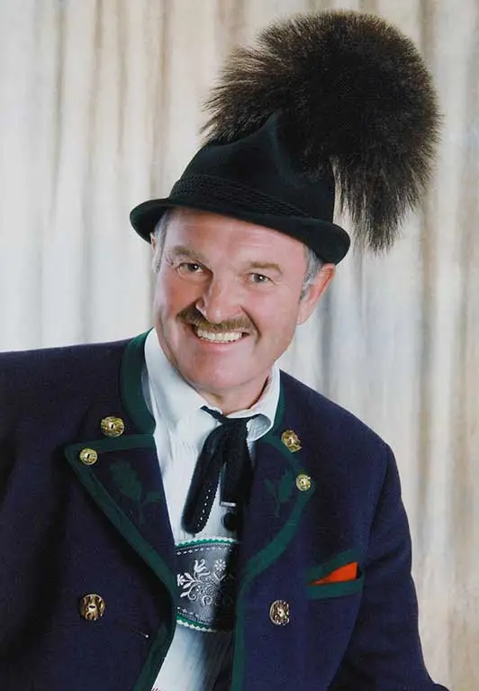 Ein Mann in traditioneller bayerischer Kleidung, darunter eine dunkle Jacke mit Zierknöpfen und ein markanter Hut mit einer großen, flauschigen Feder. Er lächelt und steht vor einem hellen Vorhang.