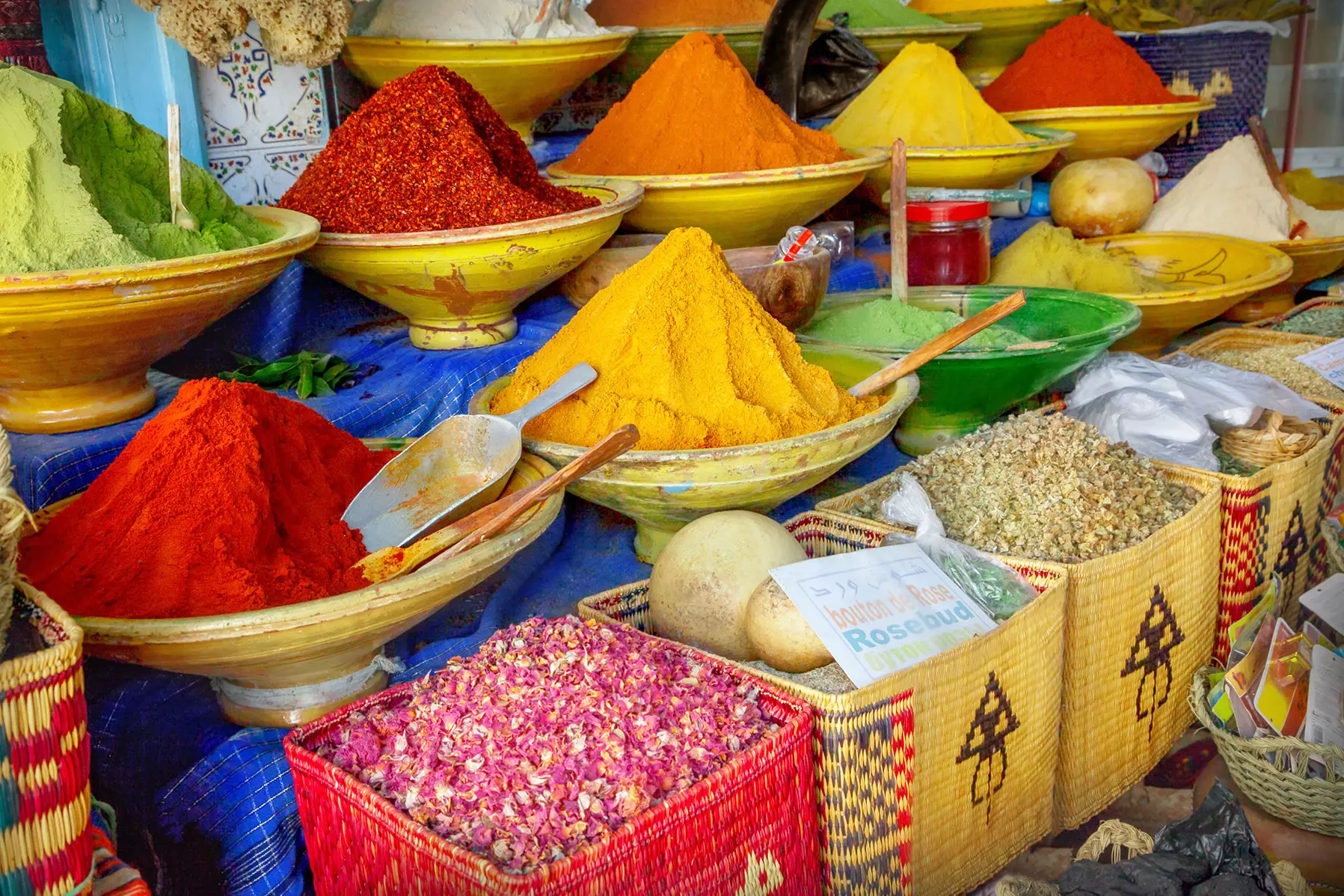 Große Schalen und Körbe mit bunten Gewürzen und getrockneten Kräutern, darunter gelbe, rote und grüne Pulver, werden an einem Marktstand mit Schaufeln und handgeschriebenen Etiketten präsentiert.