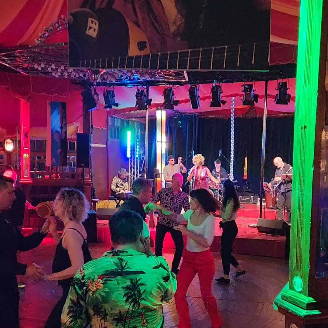 Menschen tanzen energisch vor einer Live-Band, die auf einer farbenfrohen, gut beleuchteten Bühne in einem lebhaften Lokal mit roten Vorhängen und Holzfußboden auftritt. Helle grüne und blaue Lichter erhellen die Szene.