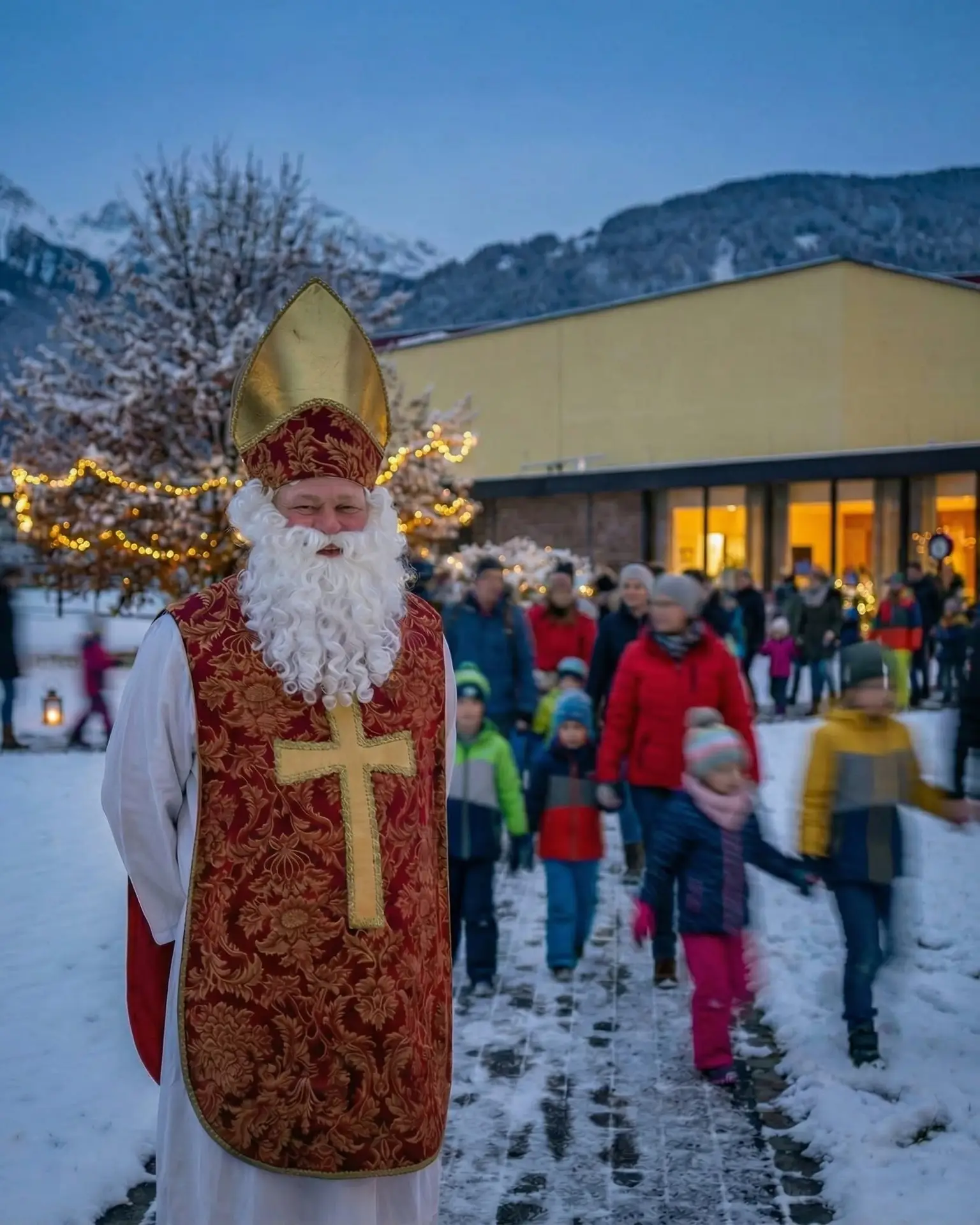 Ein als Nikolaus verkleideter Mann steht draußen im Schnee und trägt ein rotes Gewand mit goldenen Verzierungen sowie einen hohen Hut, während Kinder und Erwachsene an festlichen Lichtern und einem Gebäude in einer verschneiten Berglandschaft vorbeigehen.