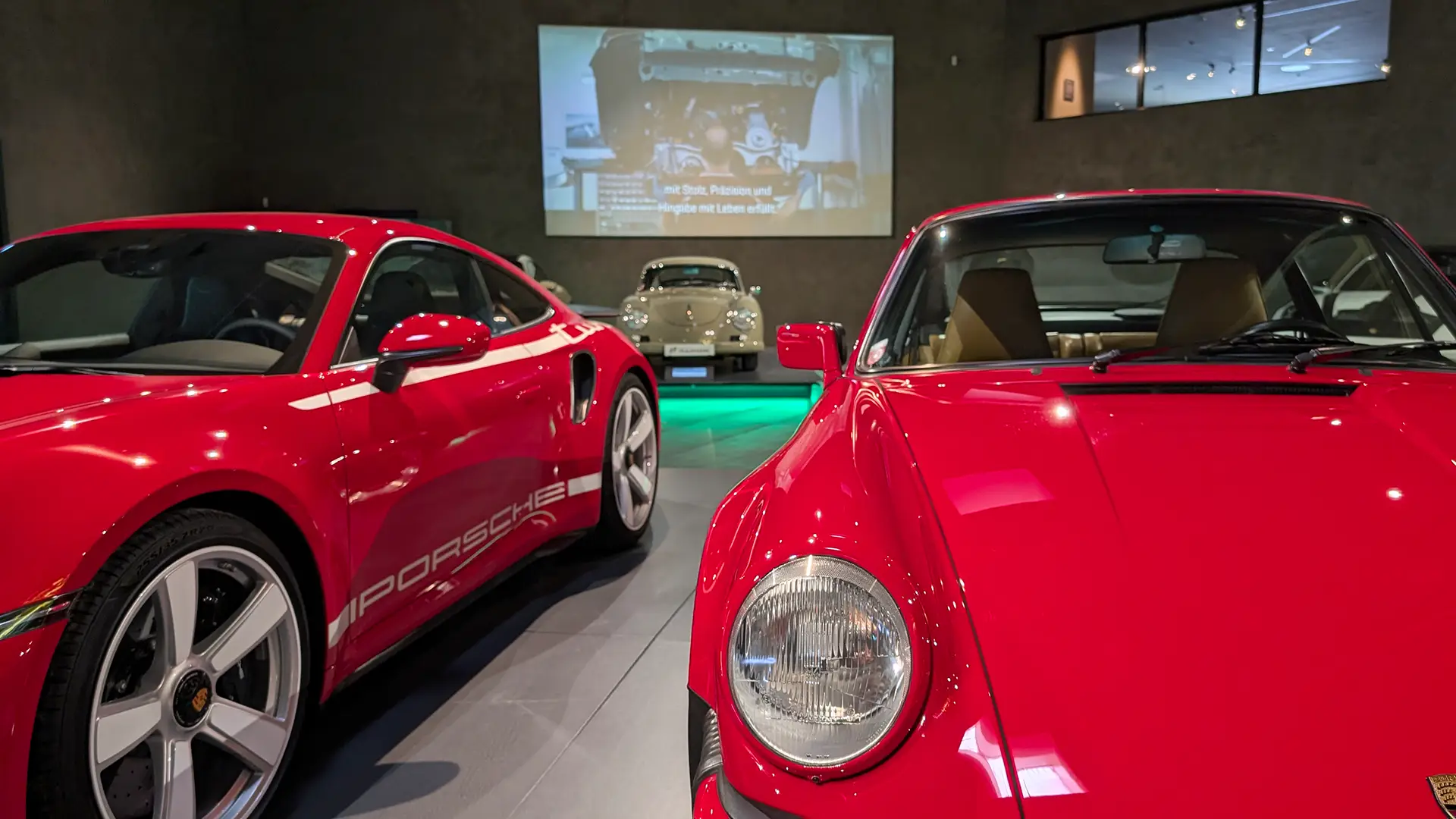 Zwei rote Porsche-Sportwagen sind im Innenraum ausgestellt, im Hintergrund steht ein klassischer silberner Porsche. An der Wand hinter den Autos wird ein Film über die Geschichte von Porsche projiziert. Das Ambiente ist modern und gut beleuchtet.