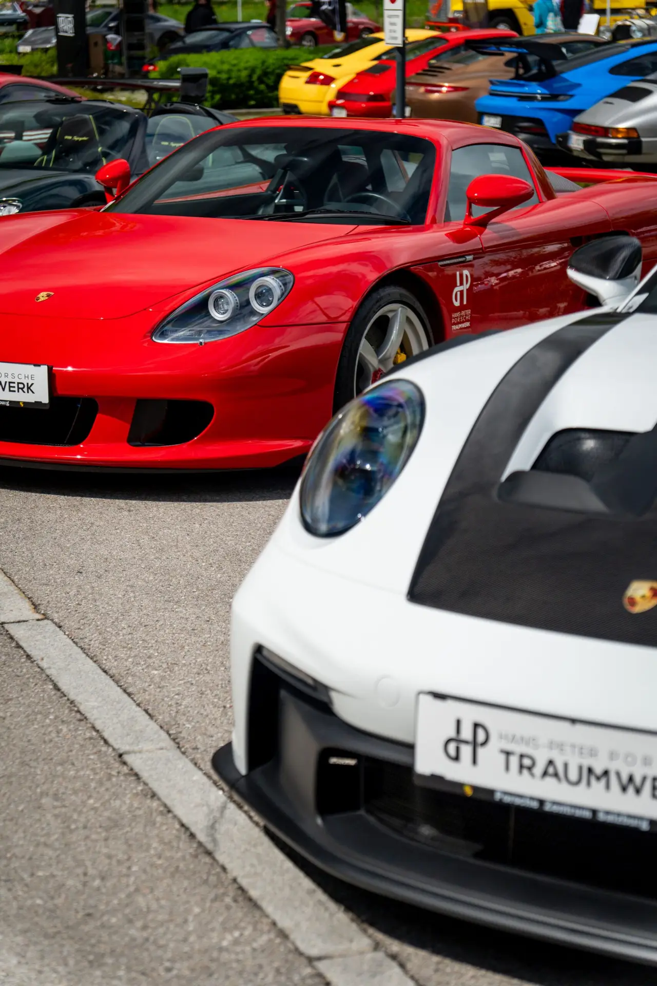 Eine Reihe luxuriöser Sportwagen, darunter ein roter Porsche Carrera GT und ein weißer Porsche, geparkt auf einer Autoausstellung mit anderen bunten Autos im Hintergrund.