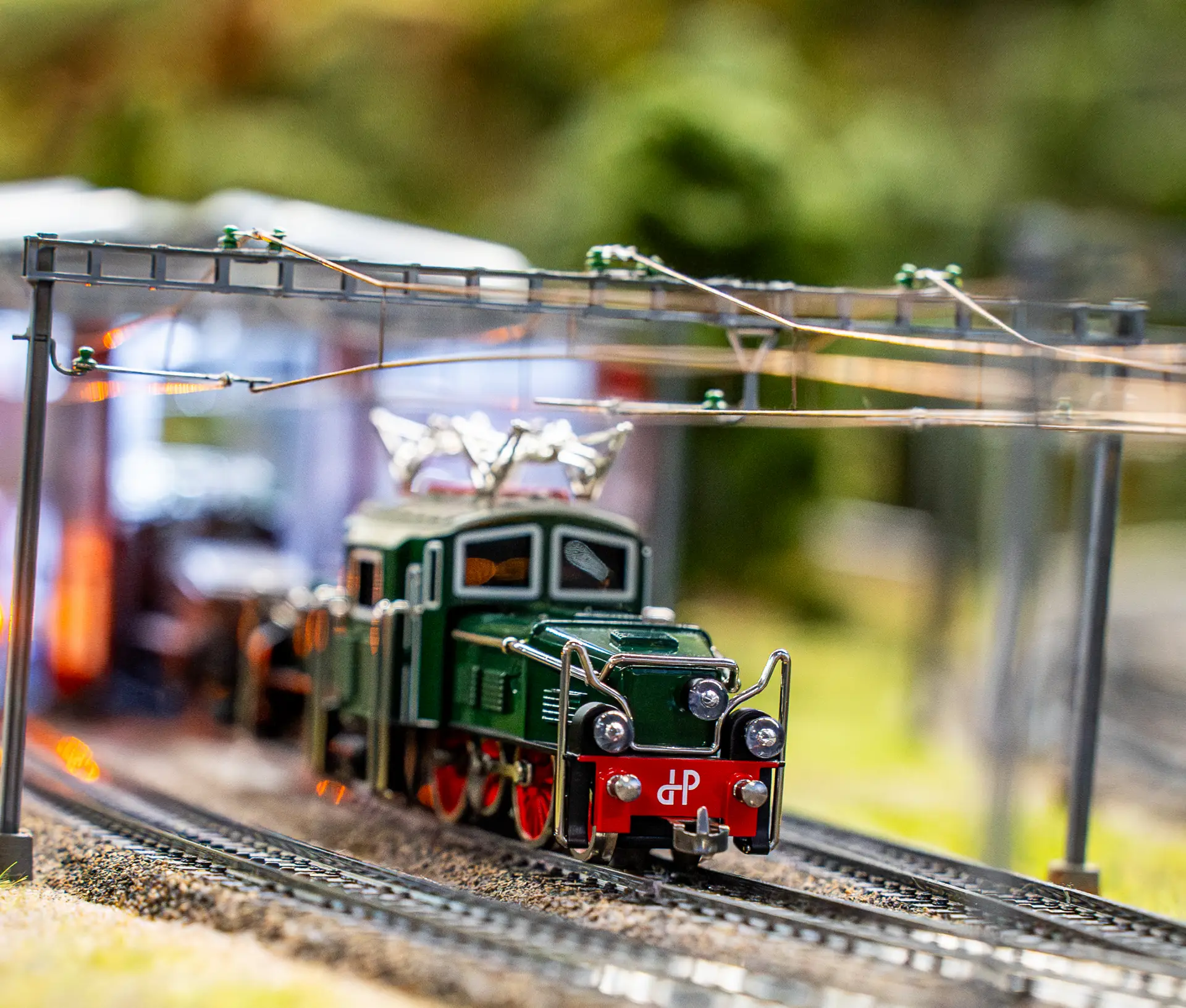 Ein detaillierter Modellzug mit grüner Lokomotive und roten Akzenten fährt auf Miniaturgleisen unter Oberleitungen, umgeben von verschwommenem Grün und Strukturen im Hintergrund.