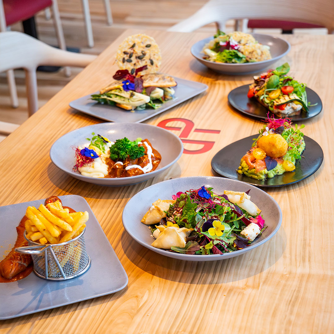 Ein Holztisch mit verschiedenen Gourmetgerichten, darunter Salate mit essbaren Blumen, Pommes frites in einem Korb und verschiedene bunte Vorspeisen auf grauen Tellern, die in einem modernen, hell erleuchteten Restaurant angeordnet sind.