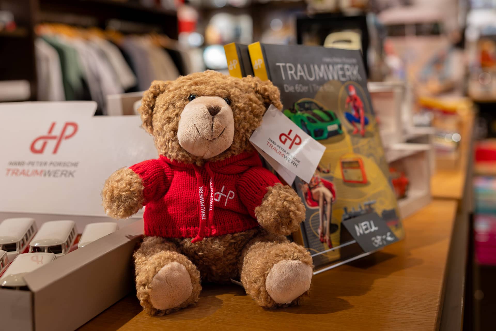 Ein brauner Teddybär in einer roten Strickjacke mit dp-Logo sitzt in einem hell erleuchteten Geschäft auf einem Holzregal neben Büchern mit dem Titel TRAUMWERK und Modellautos.