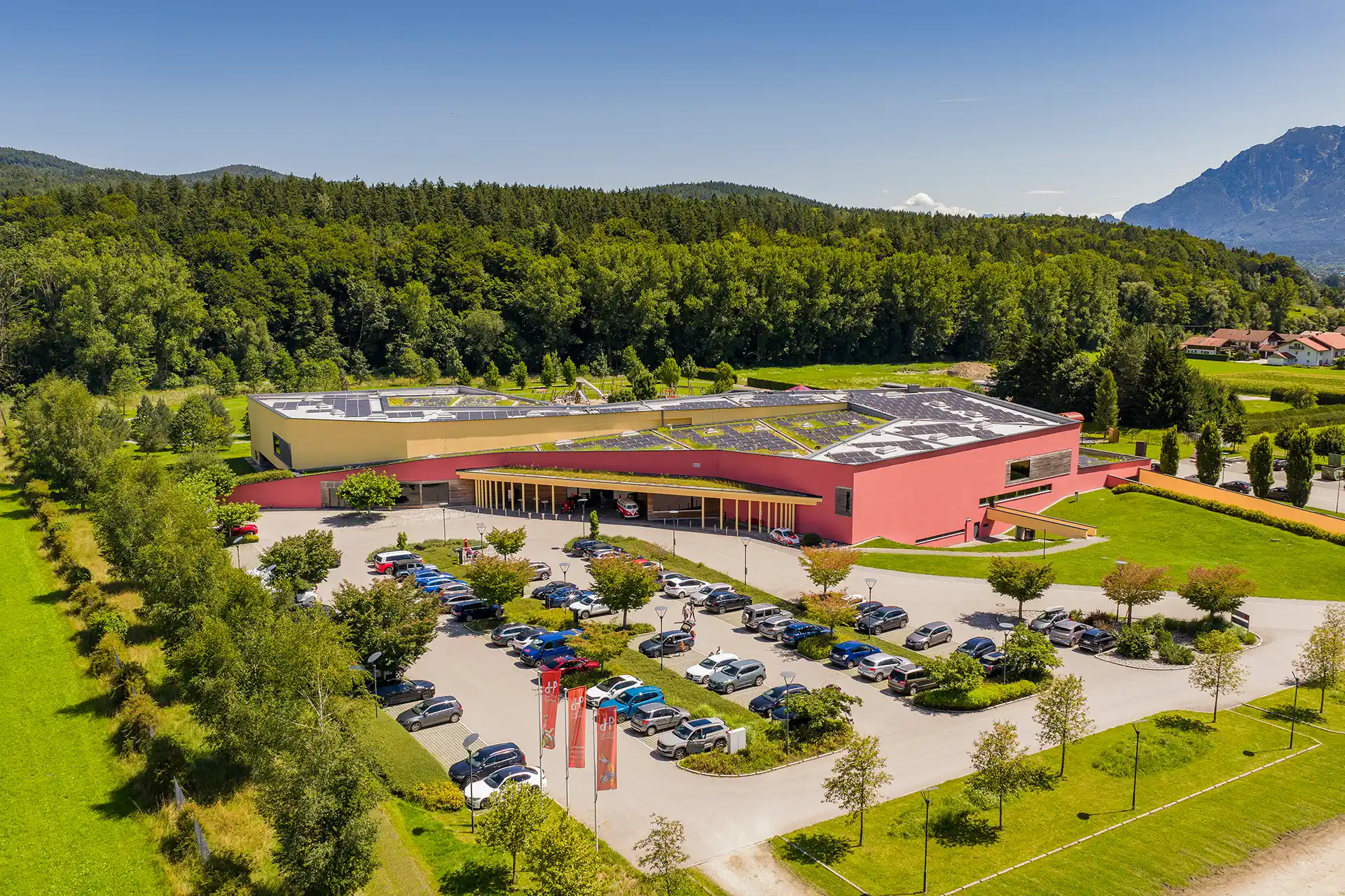Ein großes, modernes Gebäude mit rosa Fassade steht inmitten von Bäumen und Bergen. Das Dach ist mit Sonnenkollektoren bedeckt. Der Parkplatz ist voller Autos, und an einem sonnigen Tag wehen mehrere rote Fahnen in der Nähe des Eingangs.