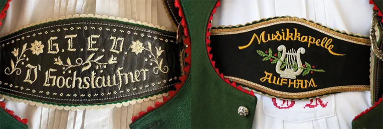 Nahaufnahme von zwei dekorativen, bestickten Abzeichen auf traditioneller bayerischer Tracht. Auf dem linken Abzeichen steht „G.T.E.V. D’Eochstaufner“ und es ist mit Blumenmotiven verziert; auf dem rechten steht „Musikkapelle Aufham“ und es ist mit einer Leier und Noten verziert.