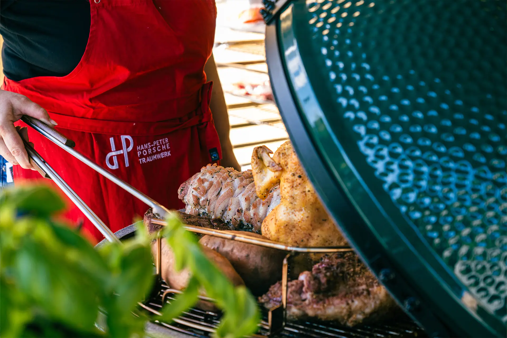 Eine Person in einer roten Schürze grillt verschiedene Fleischsorten, darunter Hähnchen, auf einem Außengrill mit grünem, gewölbtem Deckel. Im Vordergrund sind frische Kräuter zu sehen.