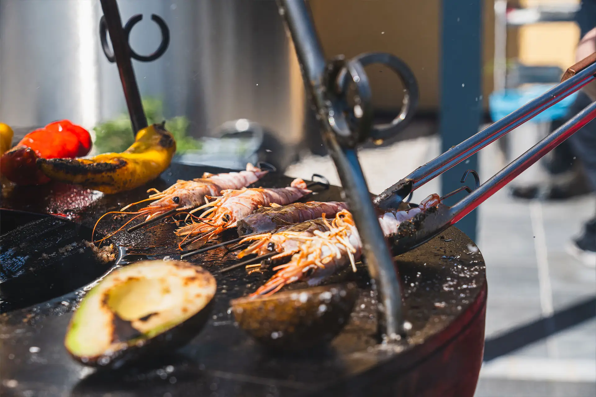 Eine Person in einer roten Schürze grillt verschiedene Fleischsorten, darunter Hähnchen, auf einem Außengrill mit grünem, gewölbtem Deckel. Im Vordergrund sind frische Kräuter zu sehen.