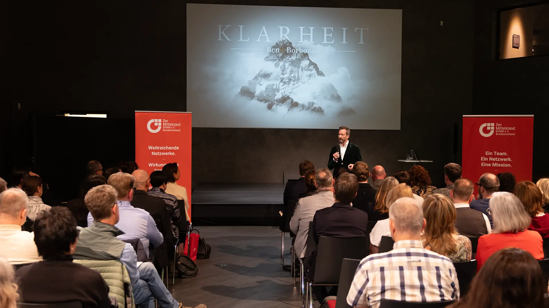 Ein Mann spricht auf der Bühne vor Publikum in einem modernen Saal. Hinter ihm ist auf einer großen Leinwand ein Berg und das Wort „KLARHEIT“ zu sehen. Auf roten Bannern zu beiden Seiten sind das Logo und Slogans des Christlichen Jugenddorfwerks Deutschlands abgebildet.
