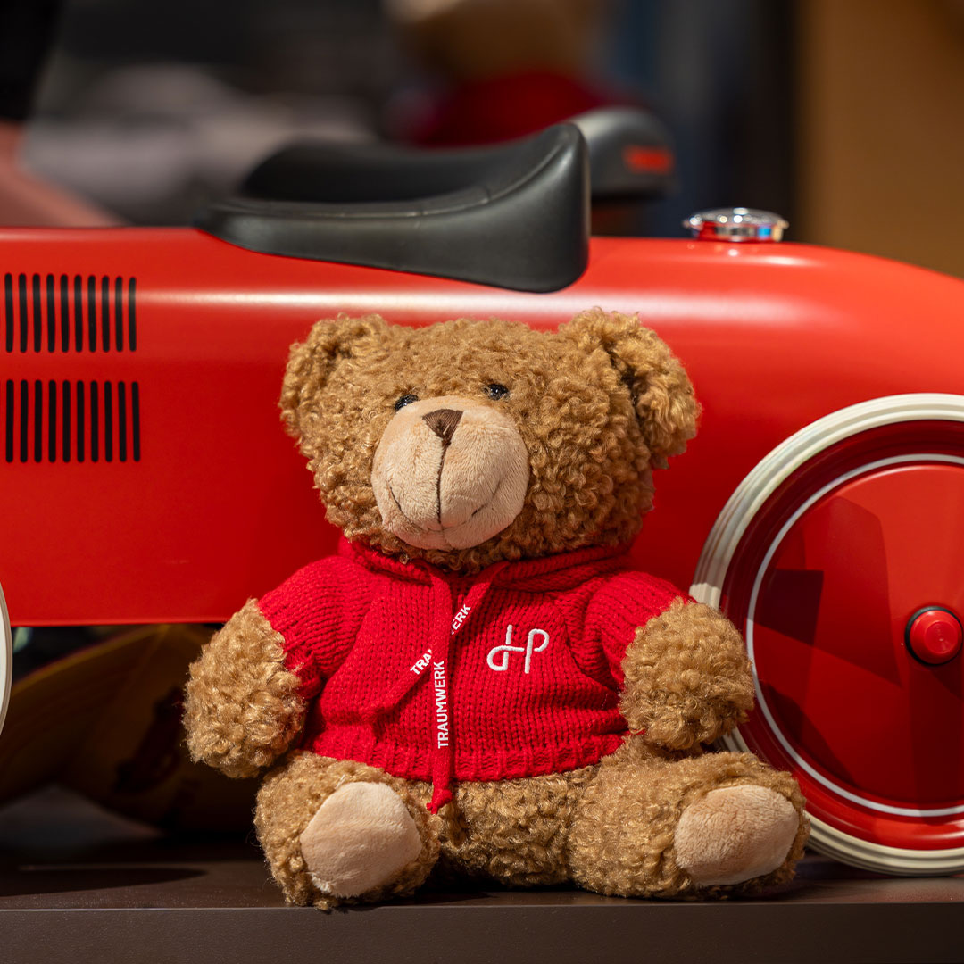 Ein brauner Teddybär, der einen roten Kapuzenpulli trägt, sitzt vor einem roten Spielzeugauto mit weißen Rädern. Der Bär befindet sich auf einer dunklen Unterlage, und der Hintergrund ist sanft unscharf.