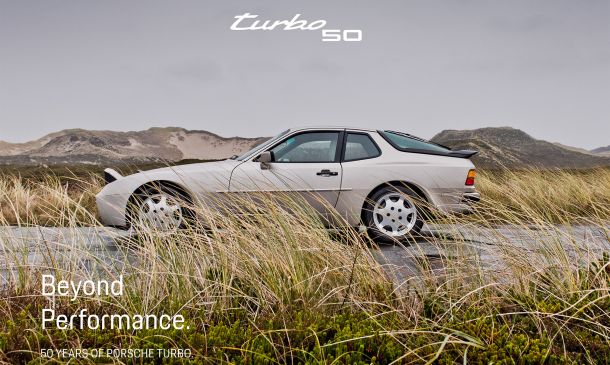 Ein klassischer silberner Porsche-Sportwagen steht auf einer Straße, umgeben von hohem Gras und Hügeln unter einem bewölkten Himmel. In weißem Text steht „Beyond Performance. 50 Jahre Porsche Turbo.“ und oben steht „Turbo 50“.