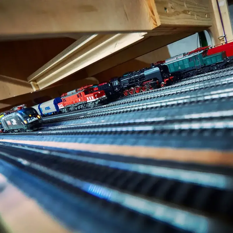 Modelleisenbahnen verschiedener Farben und Typen sind auf mehreren parallelen Gleisen unter einer Holzkonstruktion in einem Innenraum aufgereiht. Das Bild wurde aus einer niedrigen, schrägen Perspektive aufgenommen.