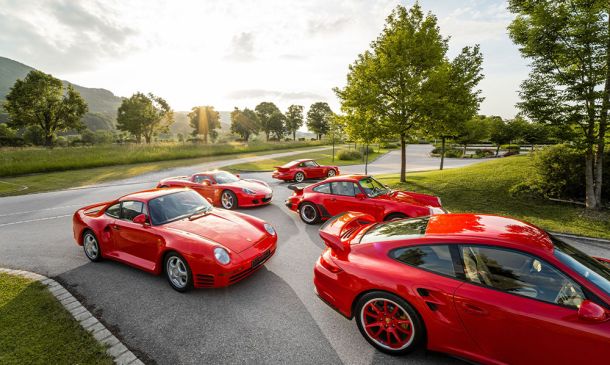Fünf rote Porsche-Sportwagen parken auf einer kurvenreichen Straße in einer malerischen, grünen Parkanlage mit Bäumen und Rasen unter einem hellen, teilweise bewölkten Himmel bei Sonnenuntergang.