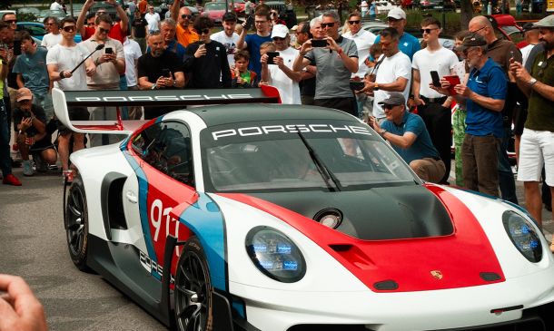 Ein Porsche-Rennwagen mit roten, blauen und schwarzen Akzenten ist bei einer Veranstaltung im Freien von einer Menschenmenge umgeben, die Fotos und Videos macht.